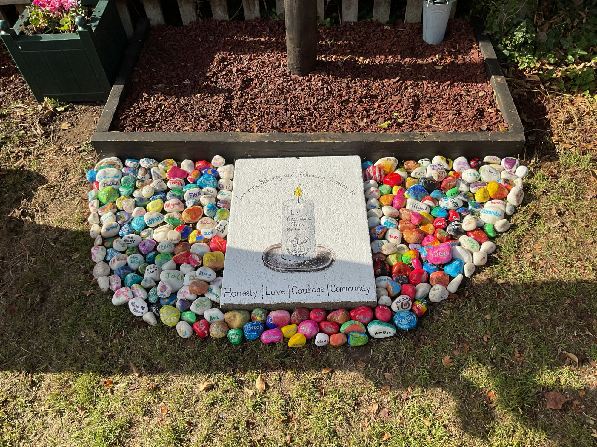 Close-up of a stone plaque featuring a candle illustration with the words “Let your light shine” and values listed as Honesty, Love, Courage, and Community. The plaque is surrounded by colorful hand-painted pebbles arranged in a heart-like shape on the ground.